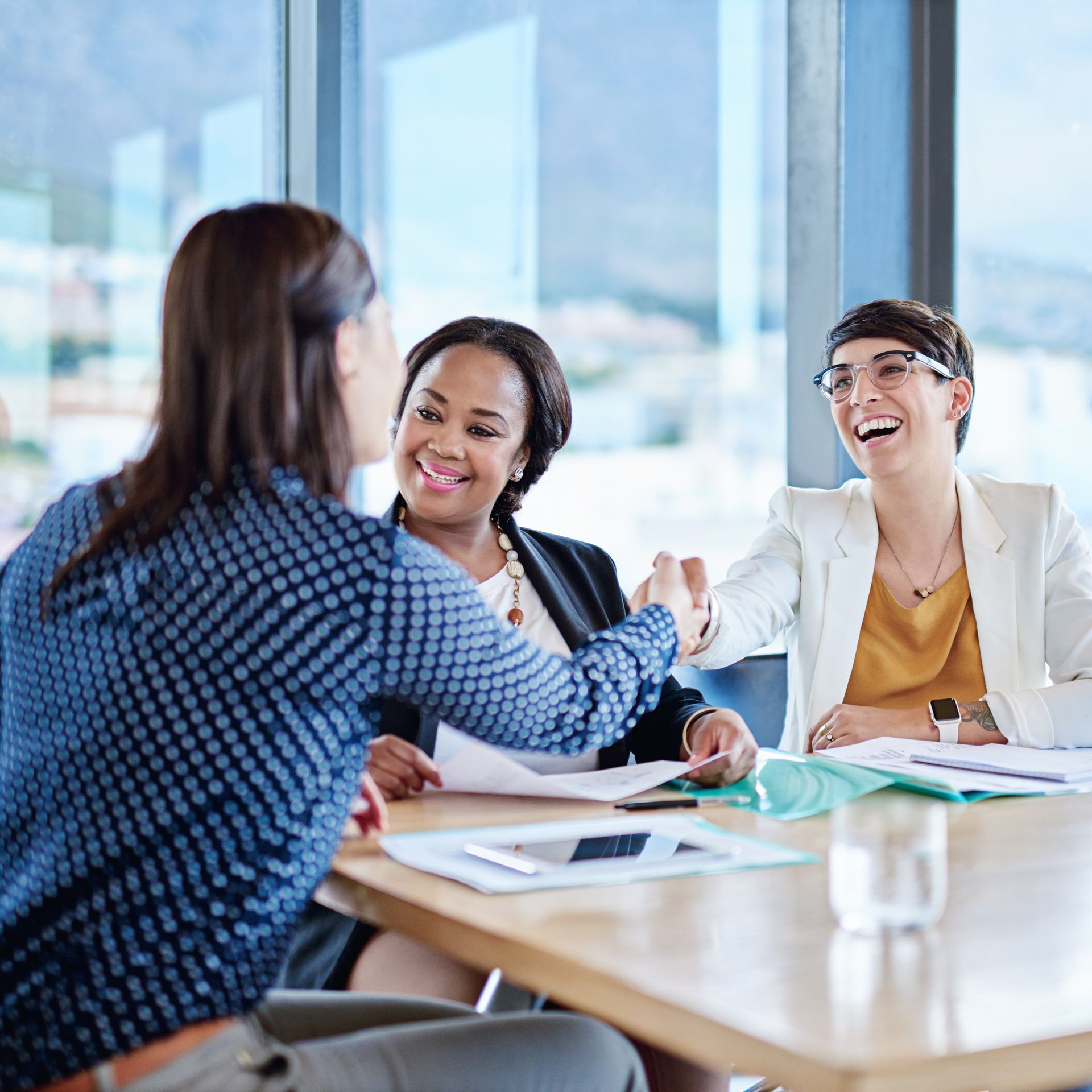 Three women in a meeting room; two shaking hands, smiling. Bright setting, sunlight, papers on the table.