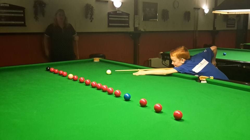 Jongen speelt snooker, mikkend op een blauwe bal omringd door rode ballen. Vrouw kijkt toe.