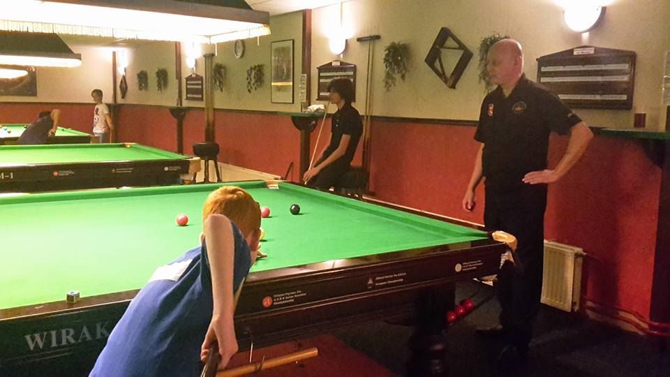 Een jongere in het blauw maakt een stootje op een pooltafel; een oudere man kijkt toe. Interieur.