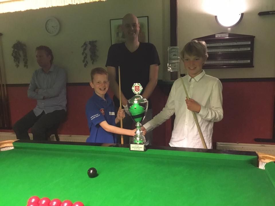 Twee jongens houden een trofee vast op een snookertafel, terwijl twee mannen toekijken.