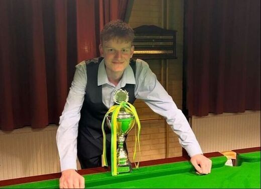 Een jongeman in een vest houdt een trofee vast op een snookertafel en lacht in een club met rode gordijnen.