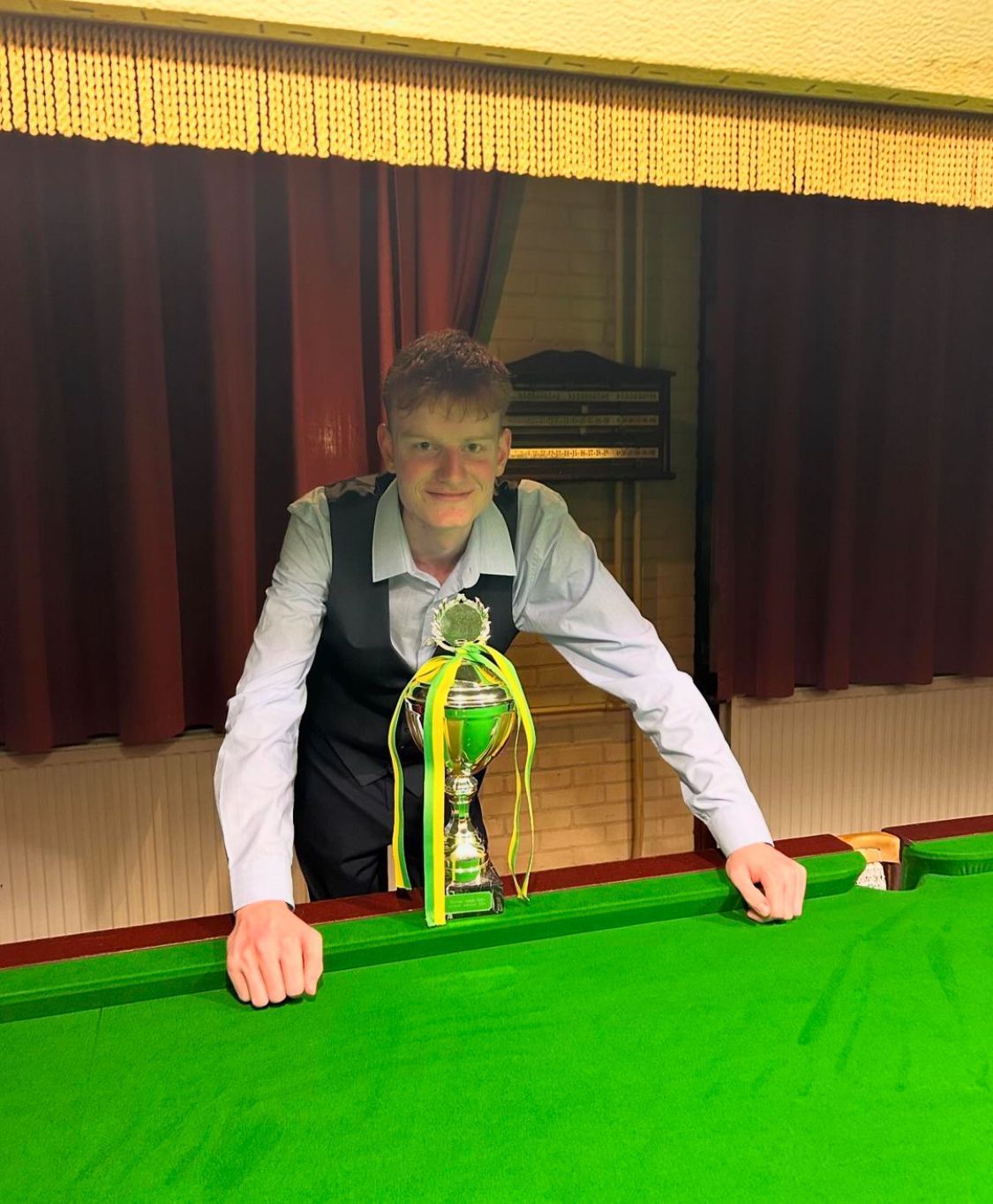 Jongeman in vest lacht, houdt een trofee vast op een groene snookertafel met een rood gordijn op de achtergrond.