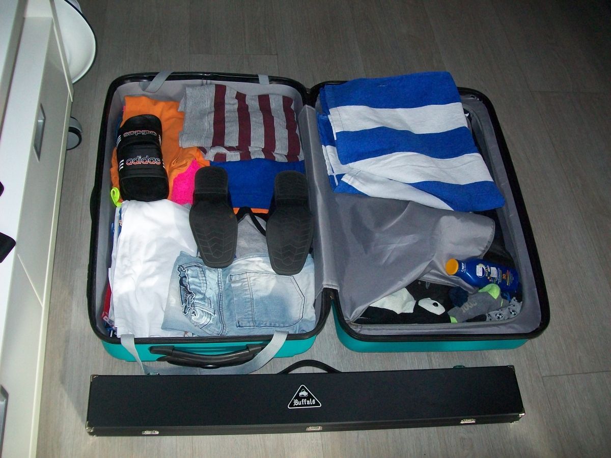 Open, blauwgroene koffer gevuld met kleding en accessoires, met een zwarte draagtas aan de voorkant.