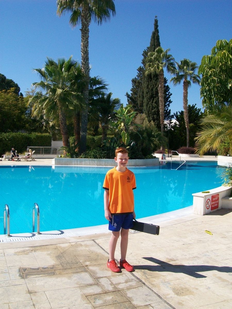 Een jongen in een oranje shirt staat op een zonnige dag bij een zwembad onder de palmbomen.