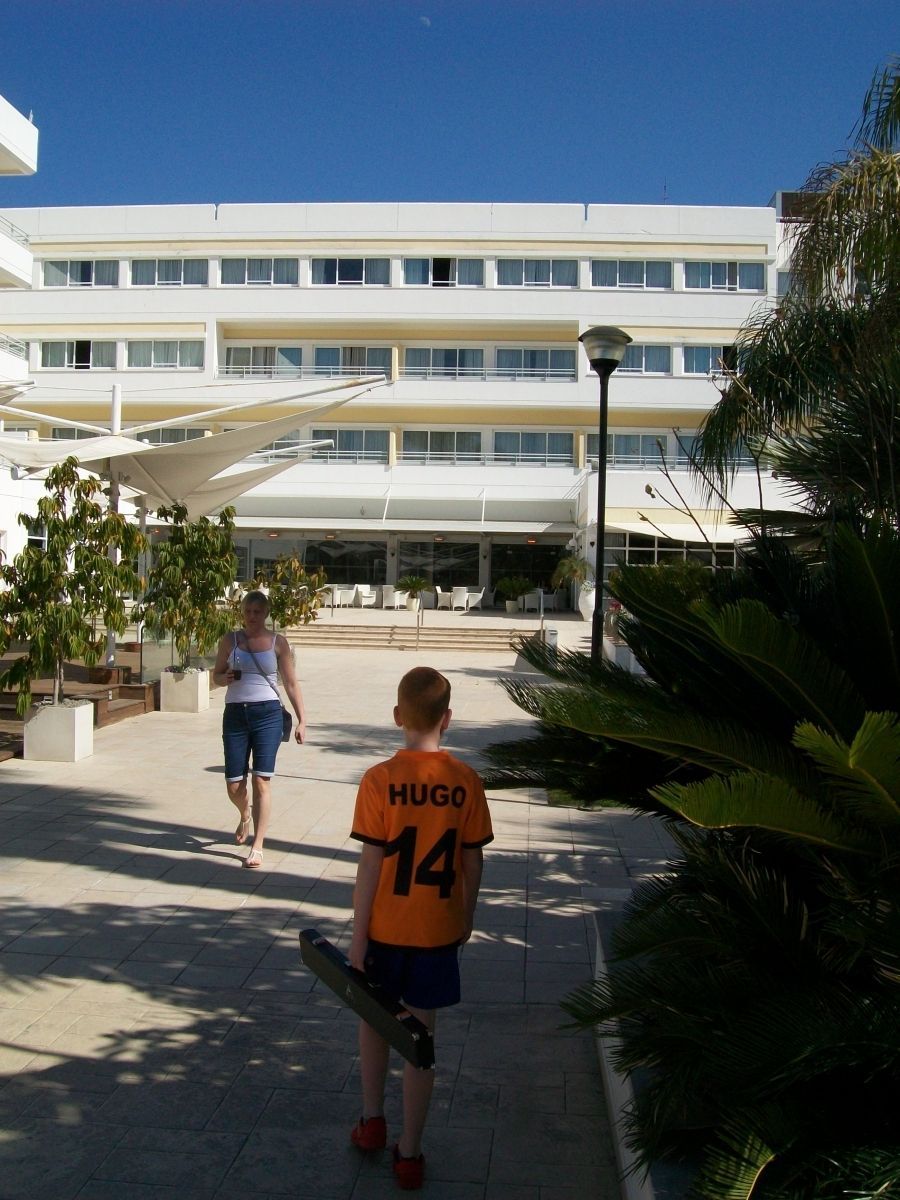 Persoon loopt naar een wit gebouw, een jongen met een skateboard. Zonnig buitenterras.