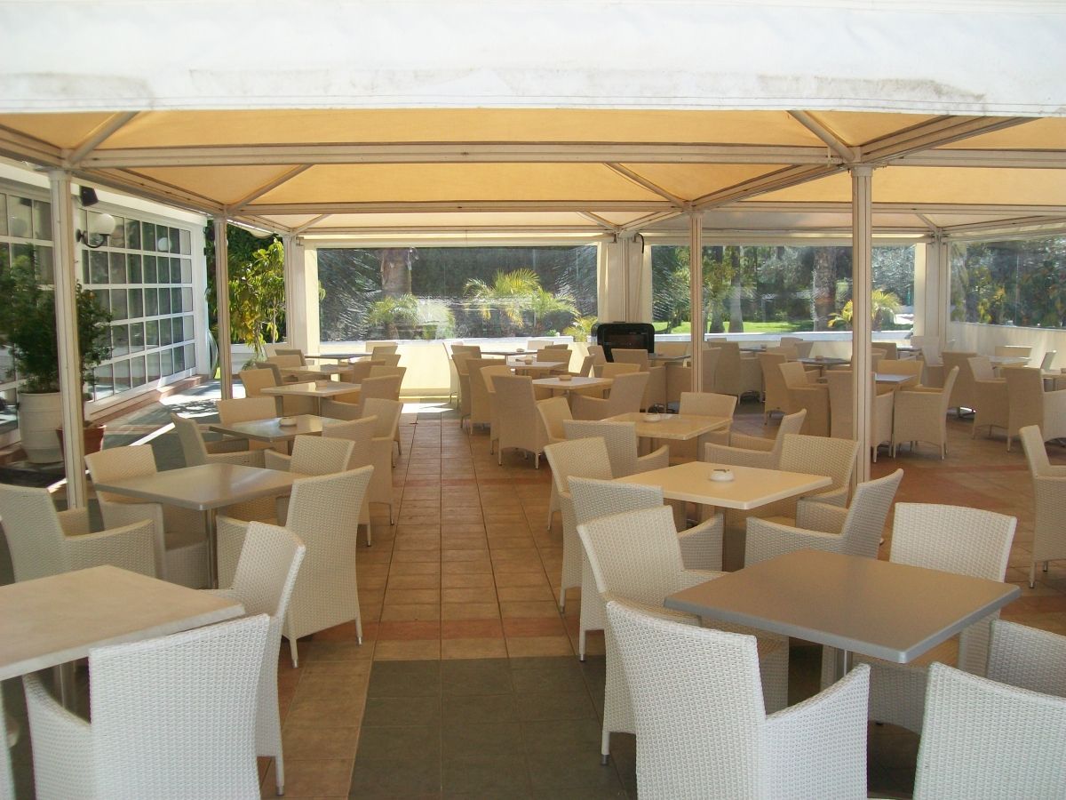 Openluchtrestaurant met witte tafels en stoelen onder een crèmekleurige luifel, met uitzicht op groen gebladerte.