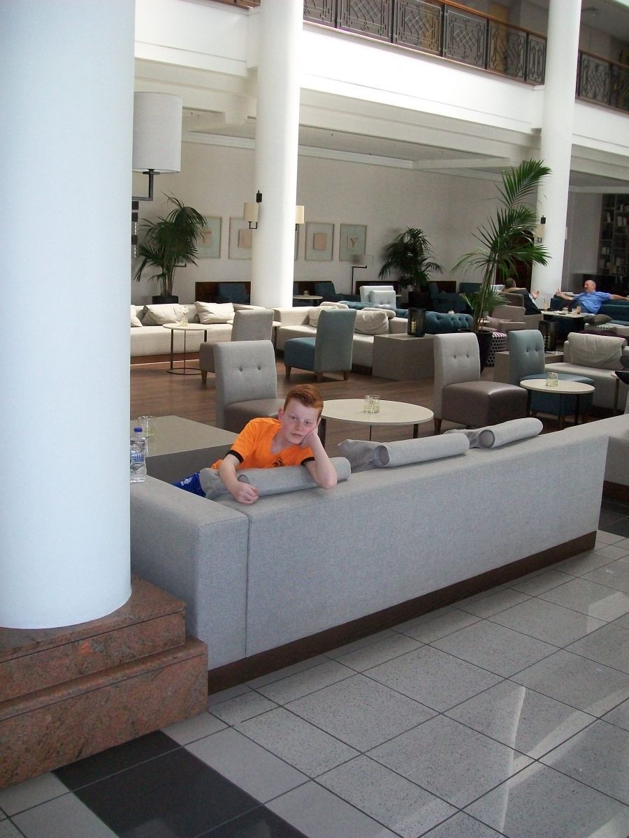 Jongen in een oranje shirt op een grote grijze bank in een hotellobby met witte pilaren en andere zitplaatsen.