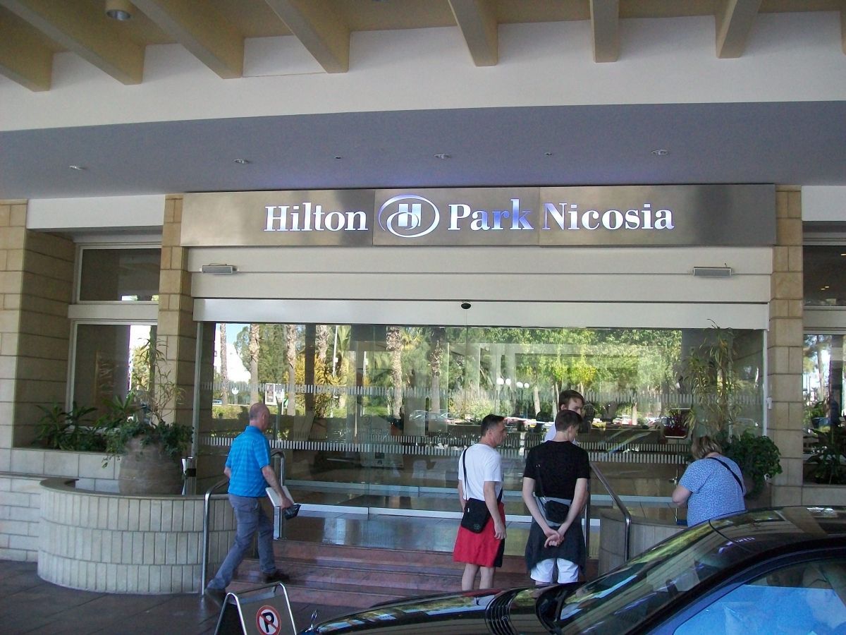 De ingang van het Hilton Park Nicosia hotel met mensen, glazen deuren en een auto.