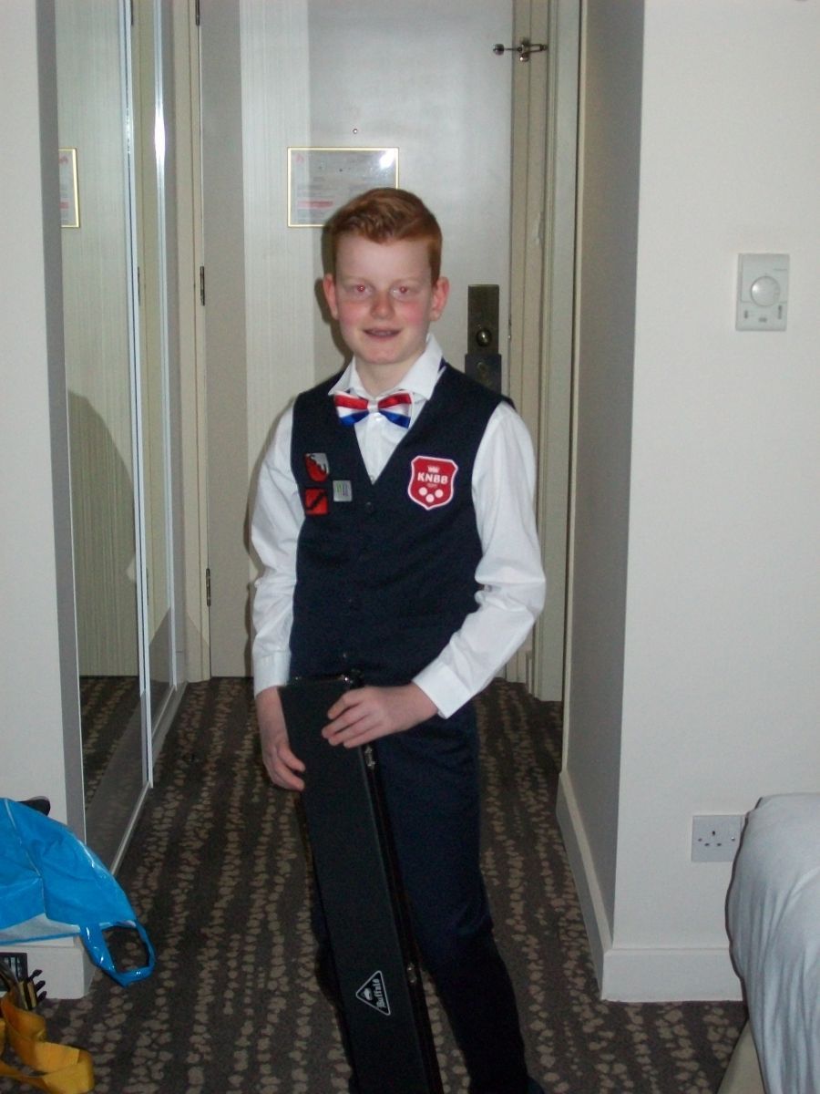 Jonge jongen met rood haar, gekleed in een vest en vlinderdas, houdt een instrumentenkoffer vast in een gang.