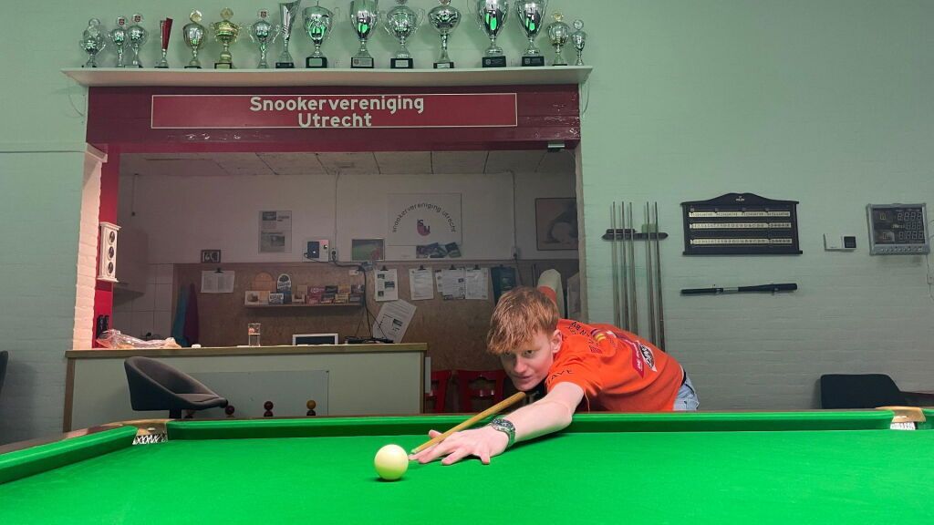 Jongen in oranje shirt speelt snooker, mikt op een speelbal, groene vilten tafel. Clubopstelling, trofeeën en borden zichtbaar.