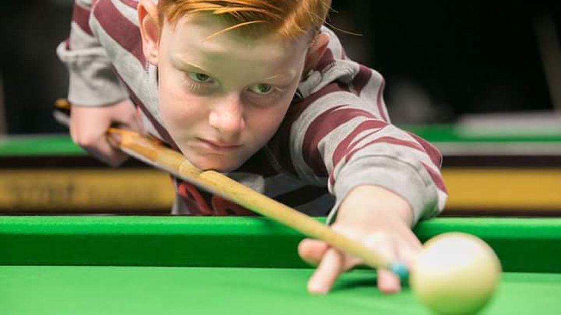Jongen met rood haar richt met een keu op de pooltafel, klaar om de witte bal op de groene tafel te slaan.