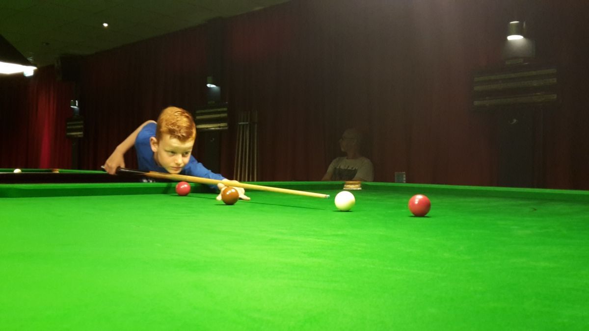 Jongen speelt snooker, mikkend op een bal op een groene vilten tafel. Rode en witte ballen zichtbaar. Donkere kamer, rode achtergrond.