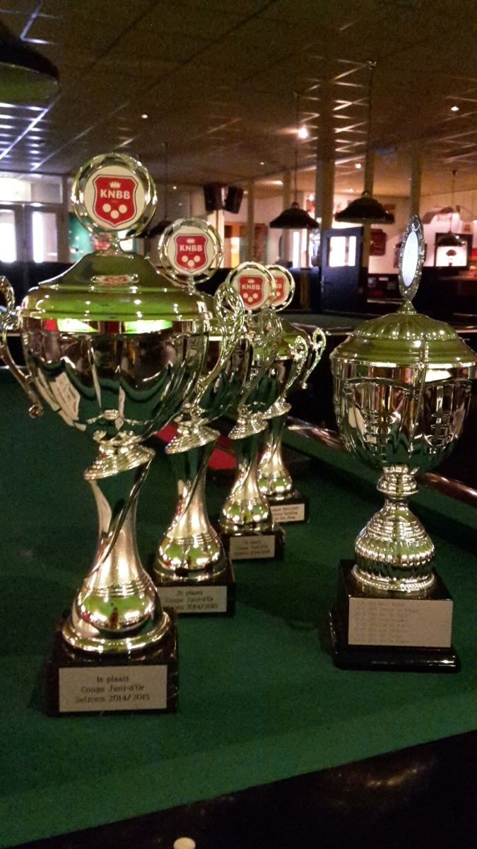 Trofeeën op een groene tafel, waarschijnlijk in een biljartzaal. Glanzend zilver met rode en witte emblemen.