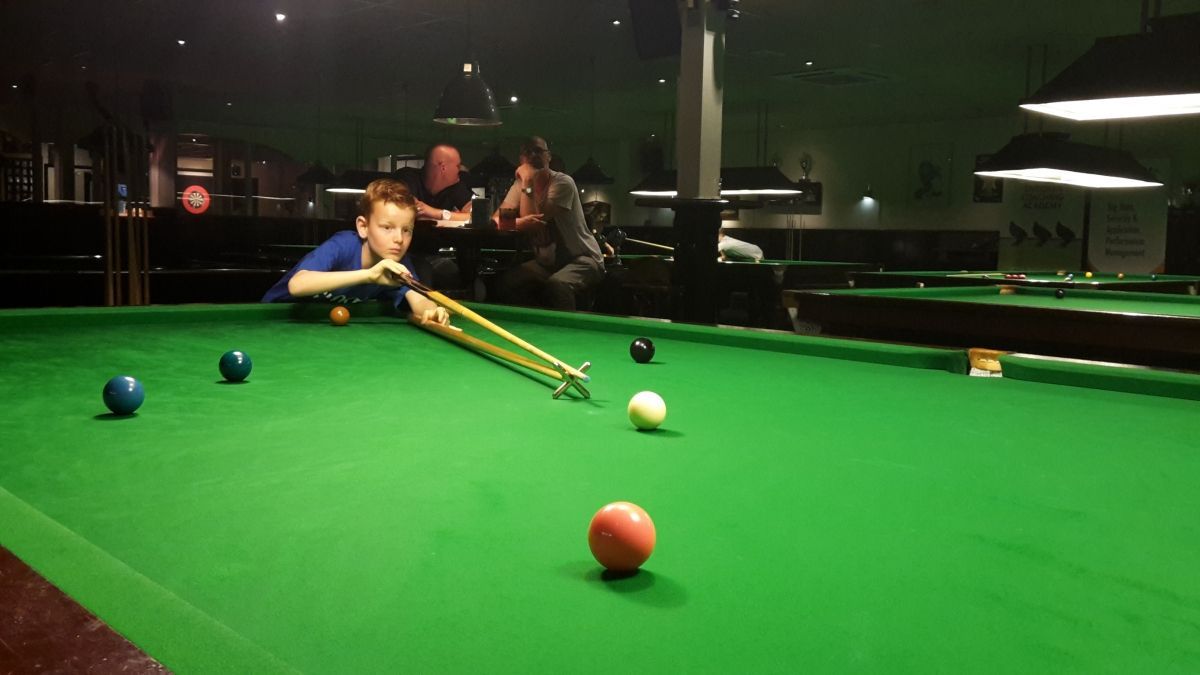 Een jongen richt een speelbal op een groene snookertafel in een schemerige snookerhal. Toeschouwers kijken op de achtergrond toe.