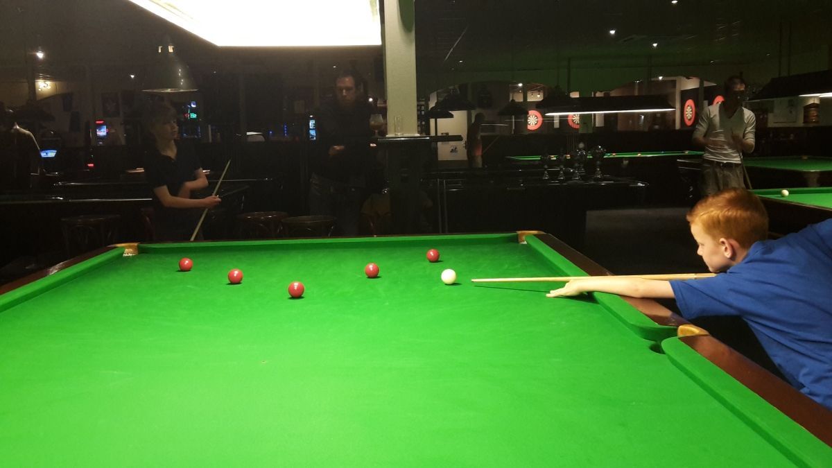 Een jongen met rood haar bereidt zijn bal voor op een groene snookertafel in een schemerige kamer, terwijl andere mensen toekijken.