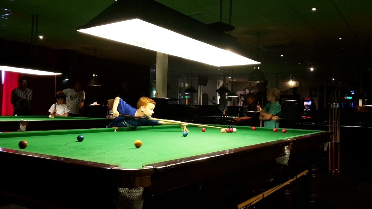 Een jongen leunt over een pooltafel en probeert een stootje te krijgen onder een felle lamp in een schemerige bar.