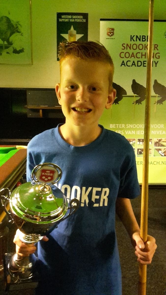 Jongen in blauw shirt met trofee en snookerkeu, lachend. Achtergrond: groene snookertafel en bord van de coachingacademie.