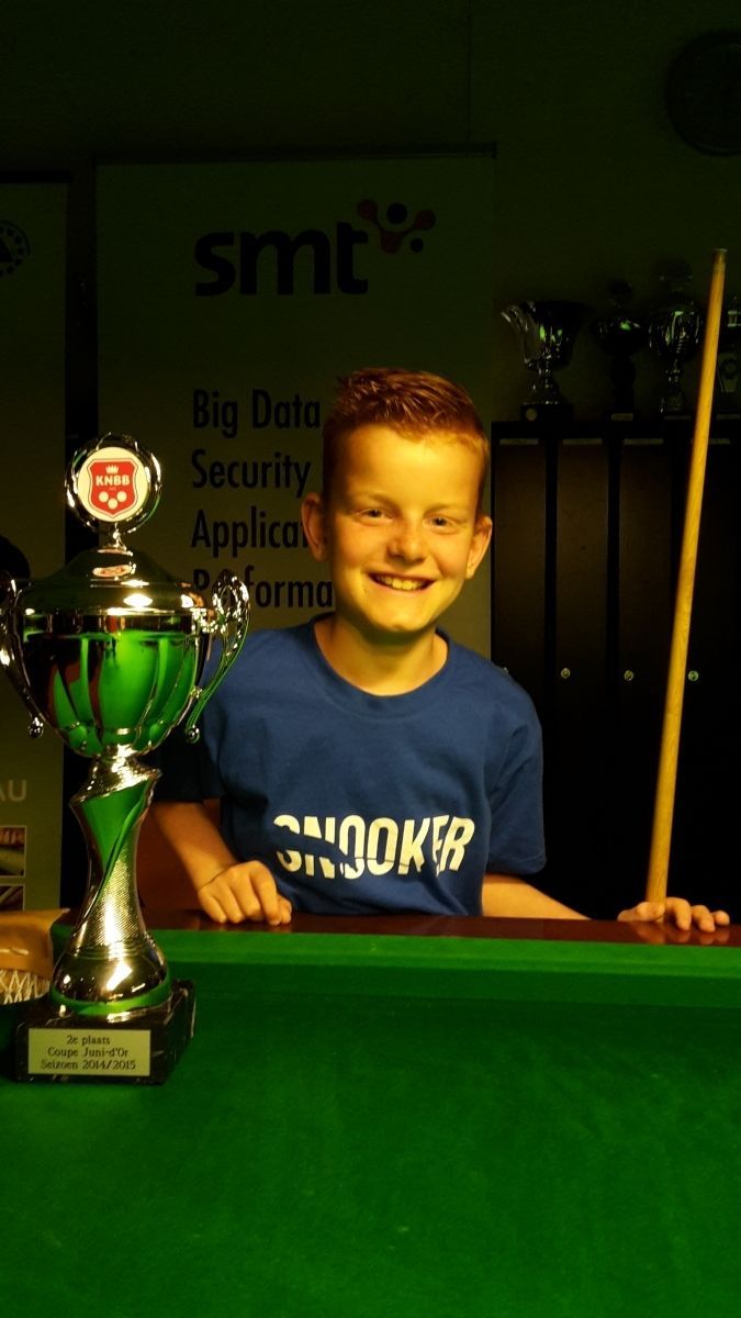 Lachende jongen met een snookerkeu in zijn hand, naast een trofee op een groene snookertafel.