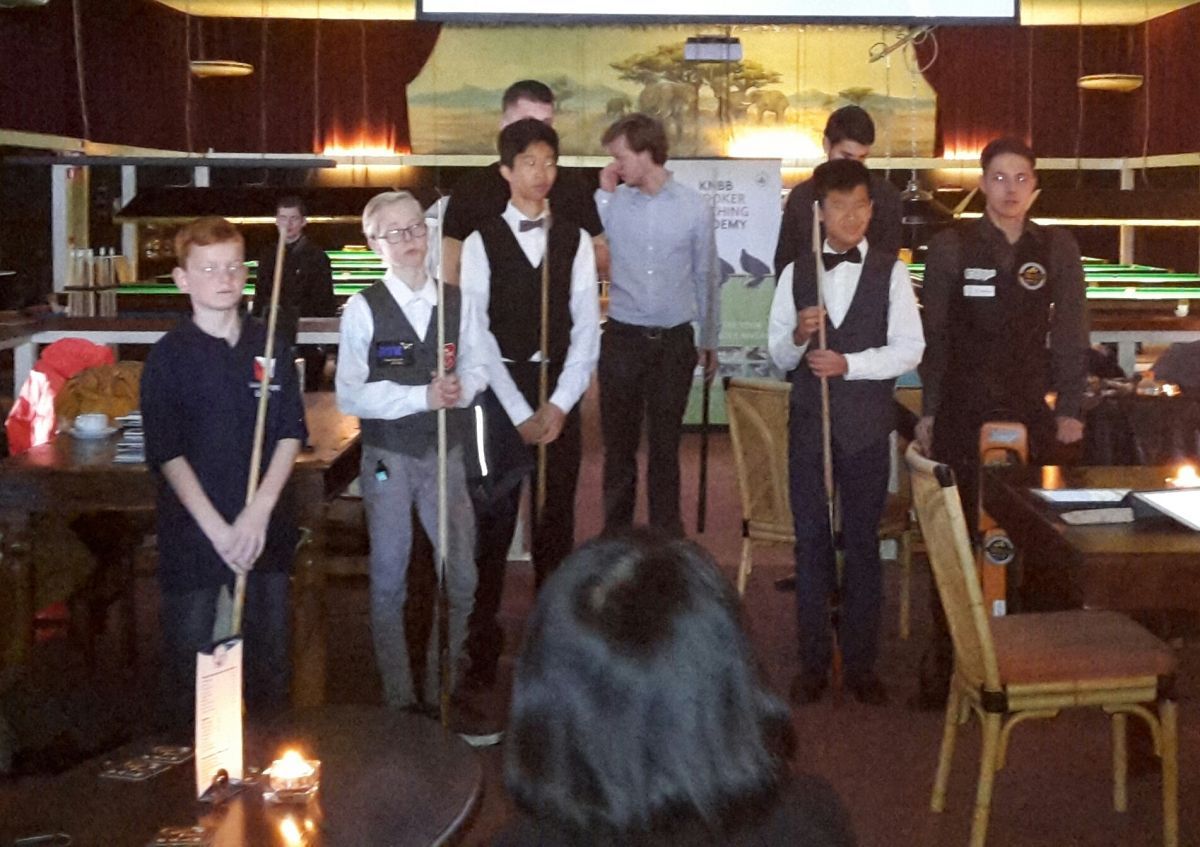 Een groep jongeren in vesten en vlinderdassen, met keuen in de hand, wachtend op hun beurt in een biljartzaal.