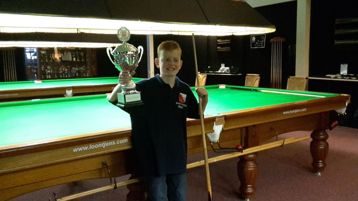 Een jonge jongen in een zwart shirt met een trofee en een keu in zijn handen, staat bij een snookertafel in een snookerhal.