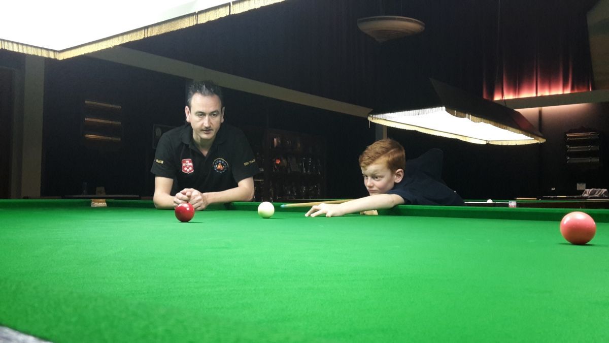 Man kijkt toe hoe een jongen een snookerballetje klaarmaakt op een groene tafel in een slecht verlichte kamer.