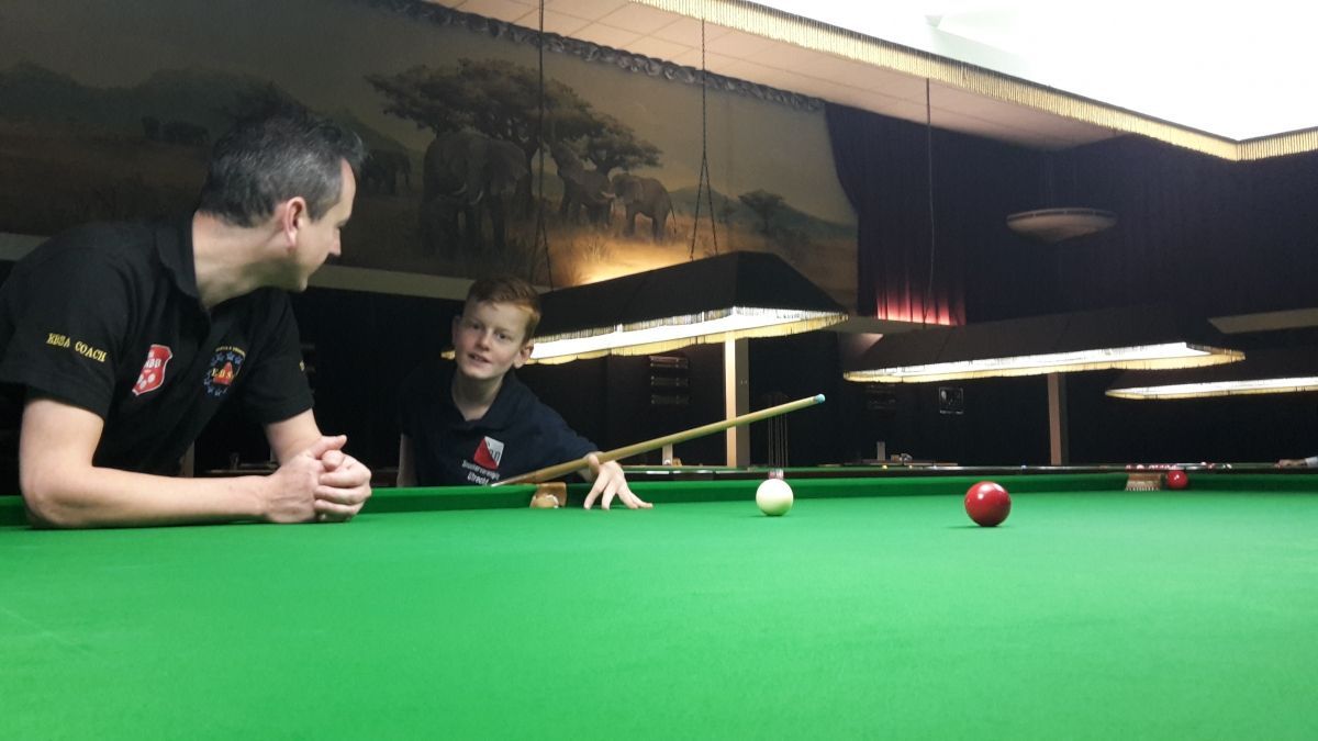 Man en kind aan een snookertafel, het kind richt, de man kijkt toe. Groene tafel, donkere kamer.