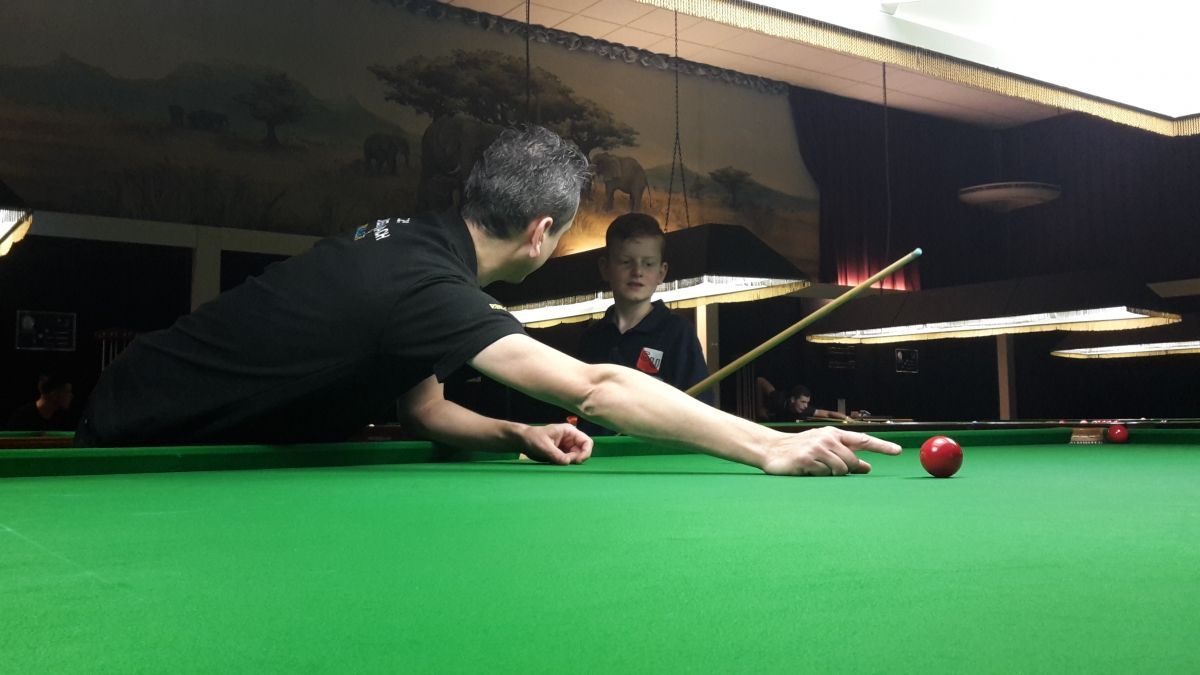 Een man bereidt zich voor op een snookertafel, terwijl iemand anders toekijkt. Groen vilt, rode bal, schemerige kamer.