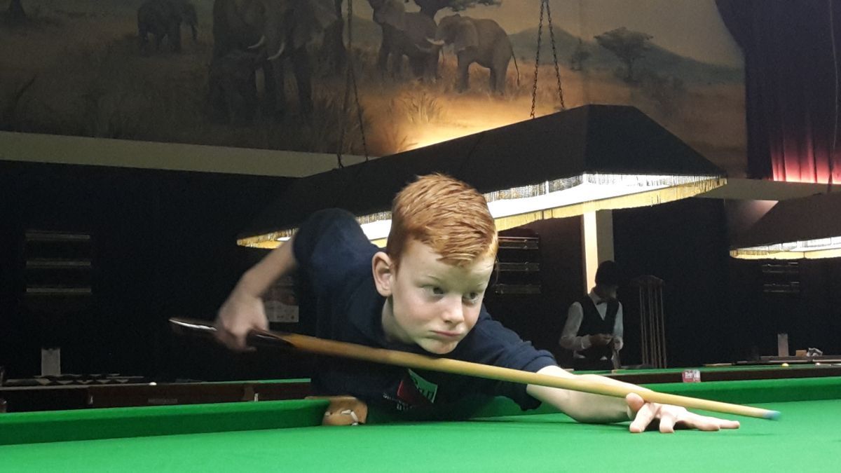 Jongen met rood haar, geconcentreerd op een groene tafel in een slecht verlichte kamer, die bezig is met het maken van een potje poolbiljart.