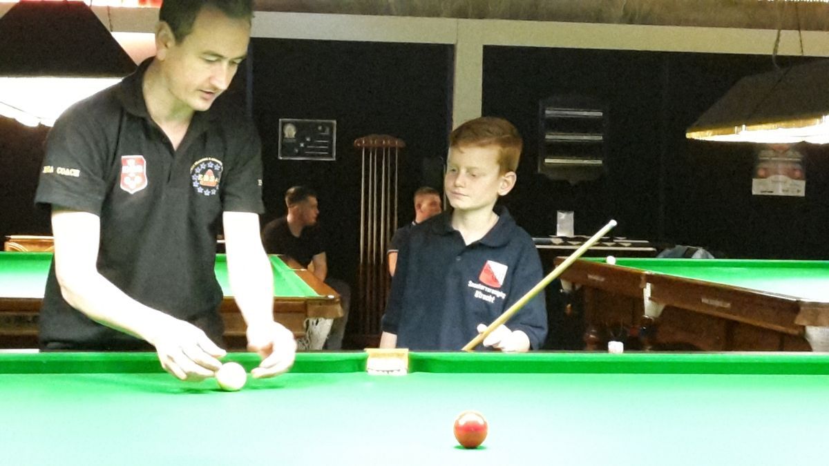 Man leert jongen snookeren, binnen, groene tafel, met ballen, keu, rode bal op tafel.