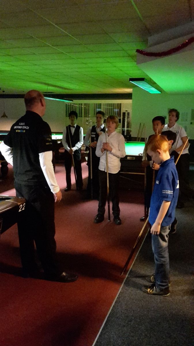 Man geeft een groep jongens les in snookeren. Donkere kamer, groen licht, jongens met keuen, rode loper.