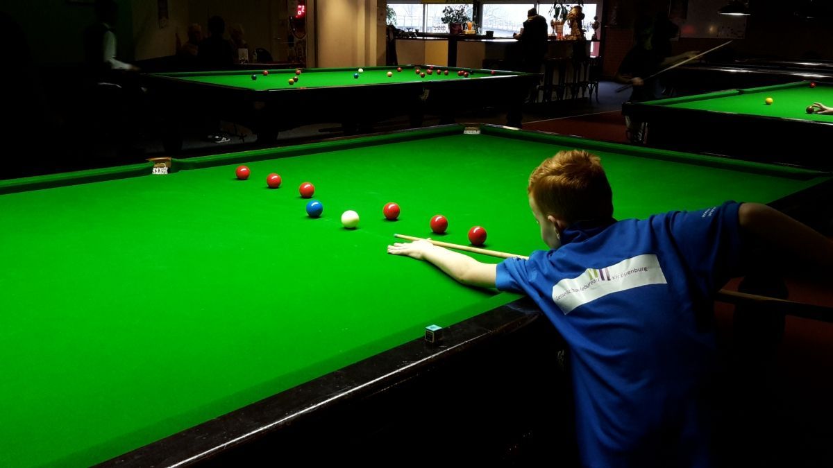 Een jonge jongen in een blauw shirt legt een bal klaar op een snookertafel, met gekleurde ballen.