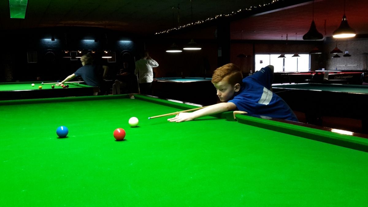 Jongen in een blauw shirt speelt snooker, mikkend op de rode bal op een groene tafel. Donker interieur.