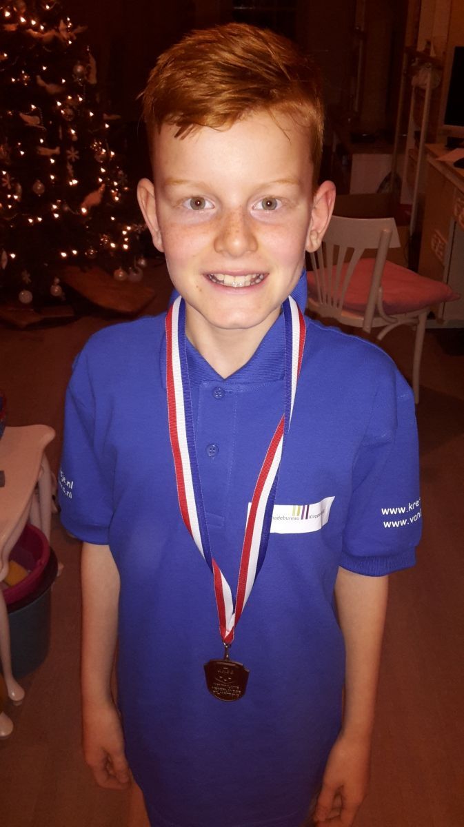 Een jongen met rood haar lacht, draagt ​​een blauw shirt en een medaille, voor een kerstboom.