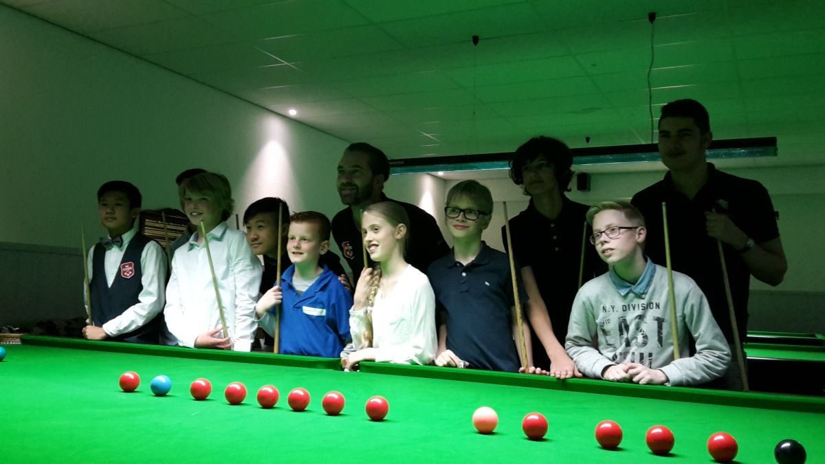 Een groep mensen, voornamelijk kinderen, rond een snookertafel. Ze kijken toe, sommigen met keuen. Groen vilt, rode en gekleurde ballen.