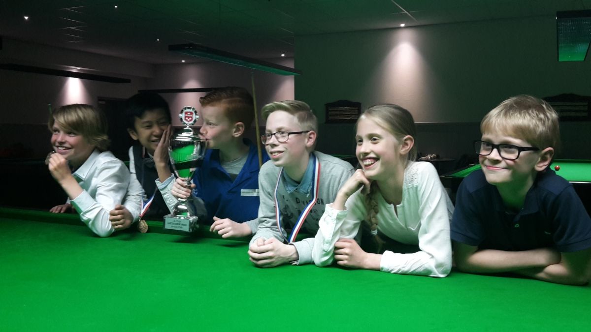 Een groepje lachende kinderen leunt op een groene pooltafel, één met een trofee in de hand, in een donkere kamer.