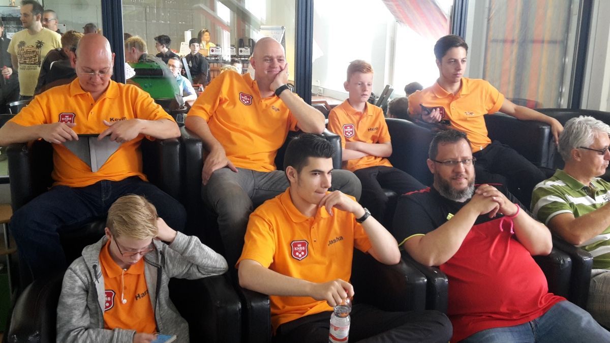 Een groep mensen, voornamelijk mannen, gekleed in oranje shirts, zittend, sommigen kijken richting de camera. Binnenlocatie.