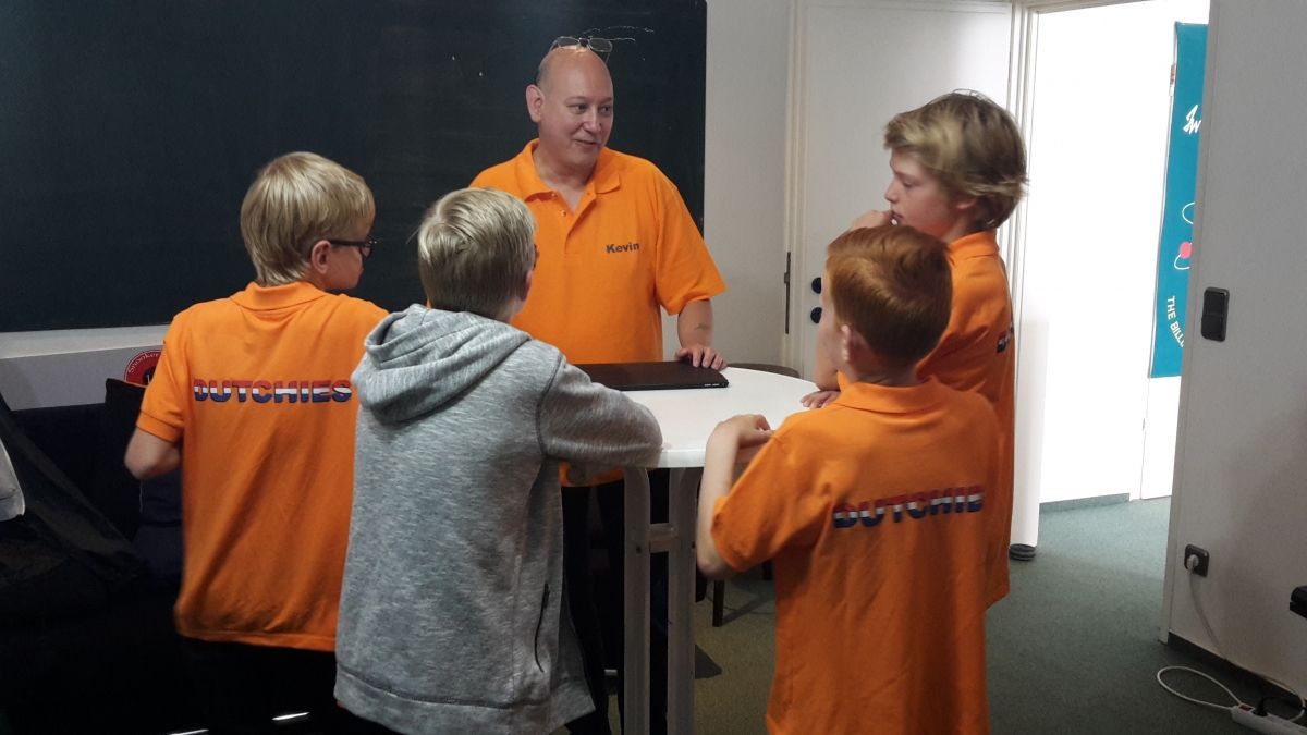 Man en vier jongens in oranje shirts zitten aan een tafel en bespreken binnen iets.