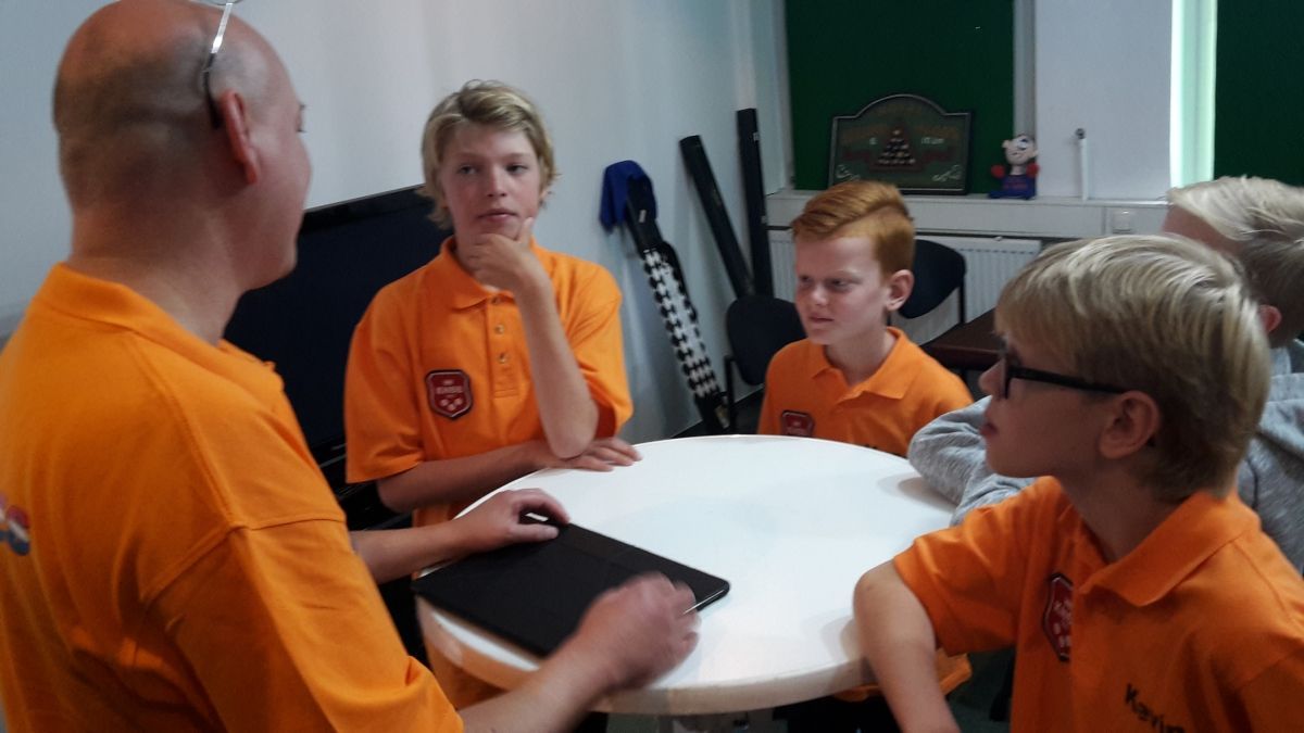 Man in oranje shirt praat met kinderen aan een witte tafel. Ze dragen oranje shirts en kijken aandachtig.
