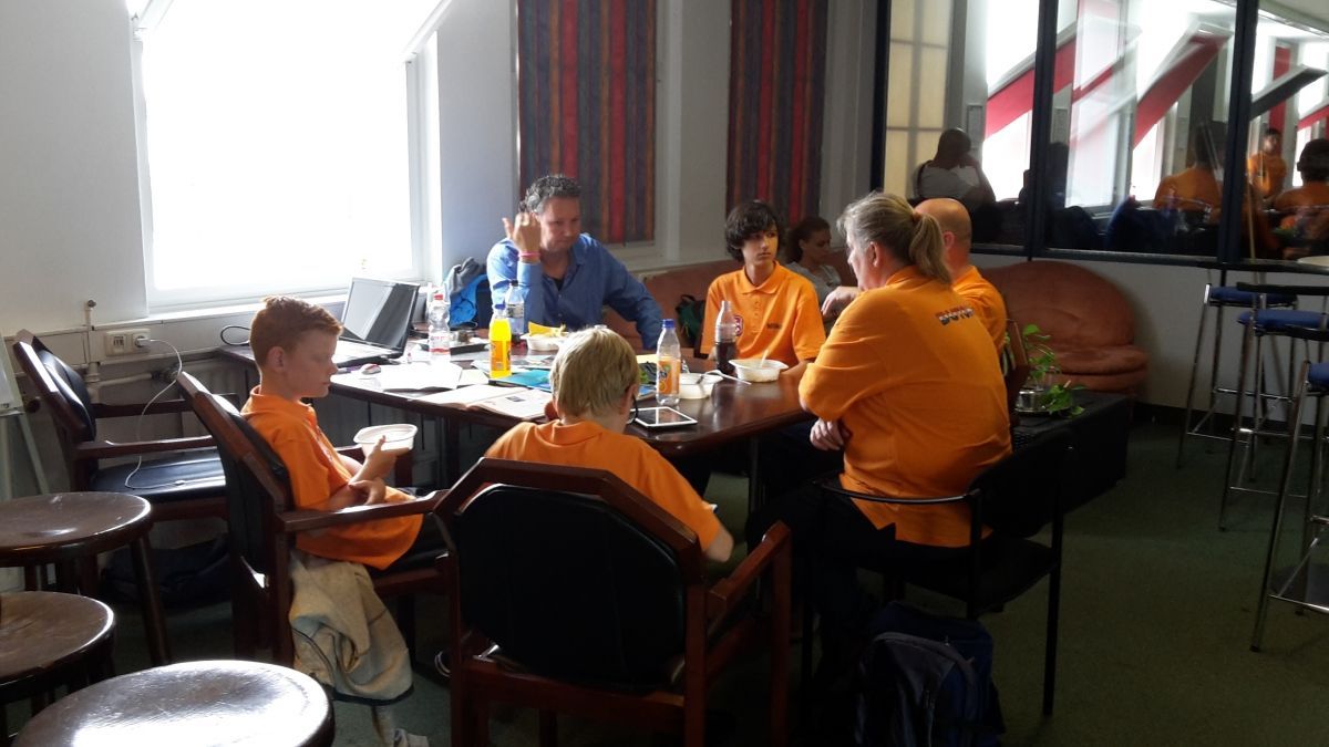 Mensen in oranje shirts en één in een blauw shirt zitten binnen rond een tafel te praten en te eten.