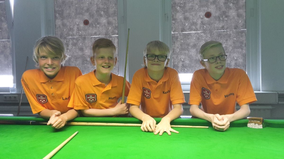 Vier jonge jongens in oranje shirts lachen, poseren achter een groene pooltafel met keu en krijt.