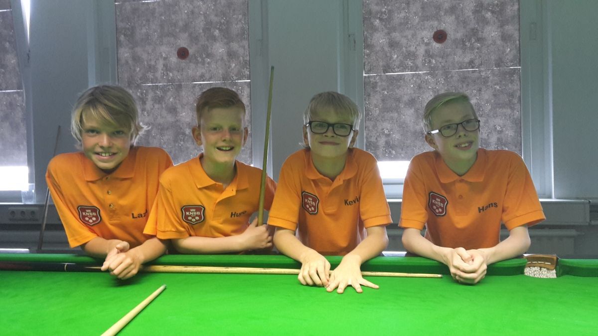Vier jonge jongens in oranje shirts lachend boven een groene pooltafel.