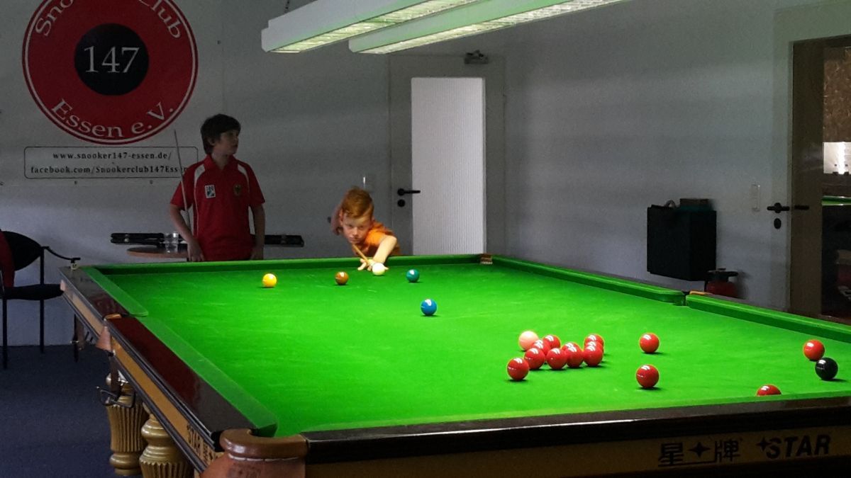 Een jong kind speelt snooker op een groene tafel; iemand in het rood kijkt toe. Binnen, lichte kleuren, ballen in zicht.