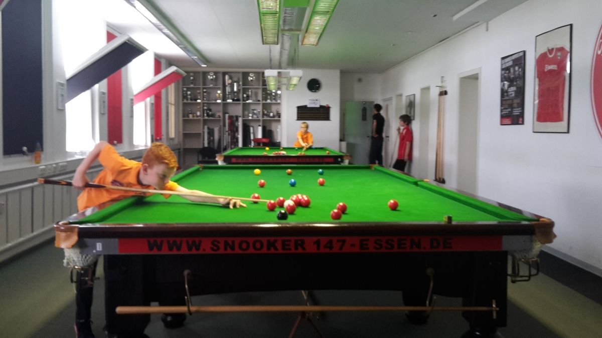 Een persoon in een oranje shirt speelt snooker en mikt op ballen op de groene tafel. Twee andere spelers kijken toe in de kamer.