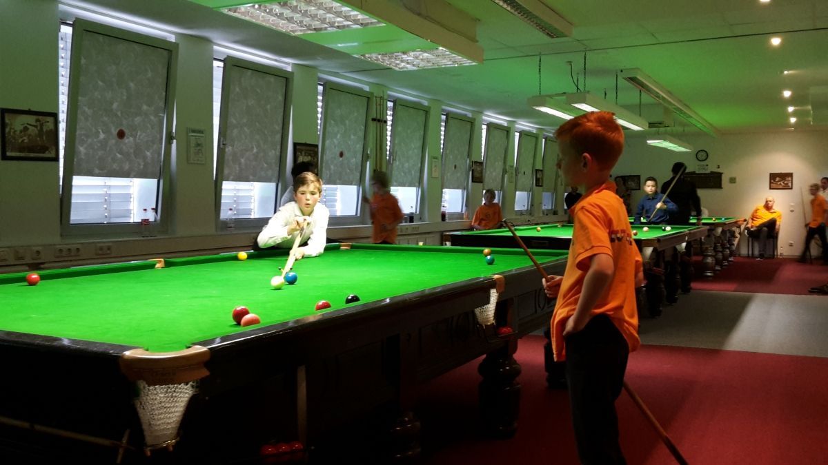 Een jongere richt met een keu op een biljartbal op een groene vilten tafel in een biljartzaal.