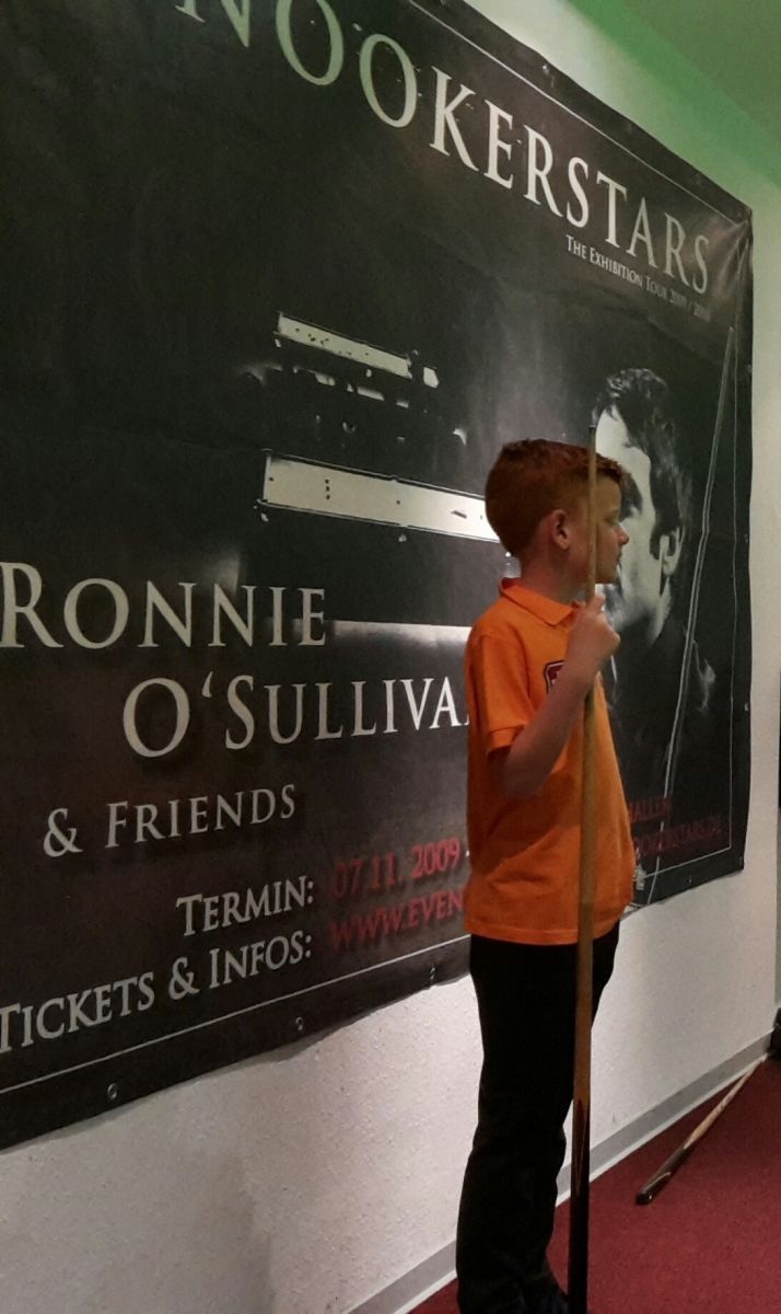 Jongen in een oranje shirt met een keu, staand voor een banner voor het snookerevenement van Ronnie O'Sullivan.