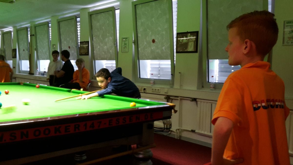 Jongen in oranje shirt kijkt naar een andere jongen die snooker speelt. Verschillende andere kinderen in een kamer met een groene vilten tafel.