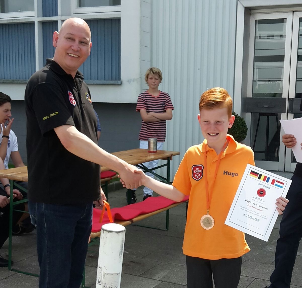 Man schudt de hand van een jongen in het oranje en ontvangt buiten een medaille en een certificaat.