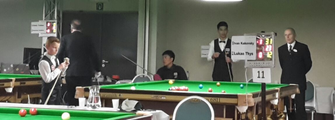 Snookerwedstrijd aan de gang; spelers en scheidsrechter rond de groene tafel.