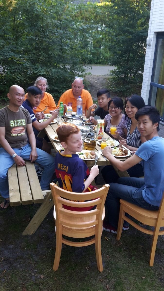 Mensen eten buiten aan een picknicktafel. Een diverse groep, een zonnige dag. Eten en drinken aanwezig.