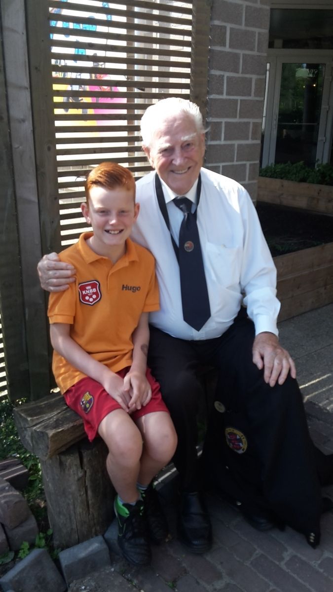Een lachende jongen met rood haar en een oudere man in uniform zitten buiten op een houten bankje.